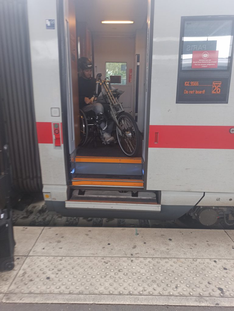 Person mit Rollstuhl und Adaptivbike im ICE, eine Zeitung lesend vor offener Tür, drei Stufen bis zum Bahnsteig