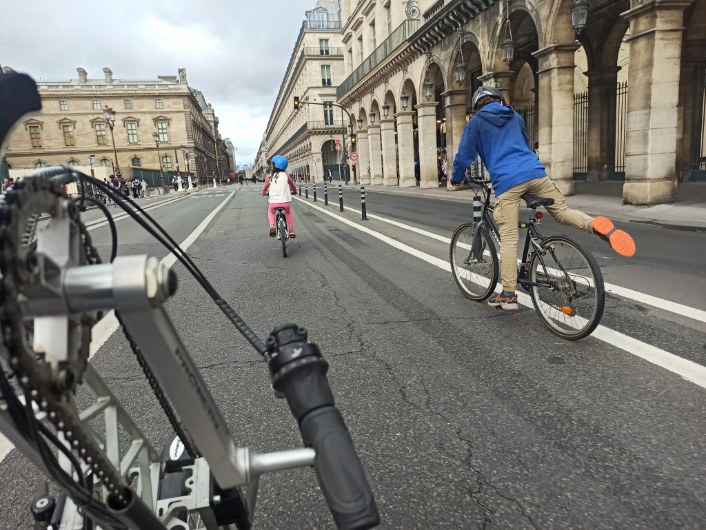 2 Kinder auf dem Rad auf einer Mittelspur einer großen Straße, die am Louvre vorbeiführt, im Anschnitt ein Adaptivbike; keine Autos